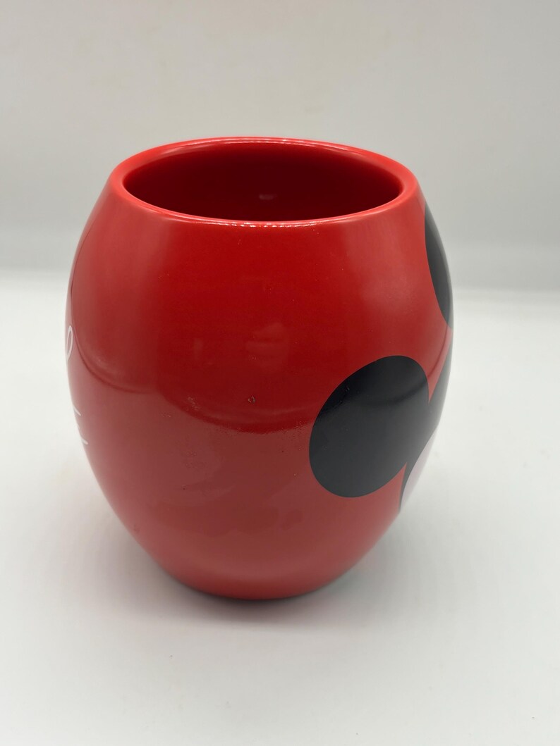 May include: A red ceramic mug with a rounded shape and a wide opening. The mug features three black circles on the side, and a white design. The mug is likely for beverages.