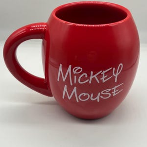 May include: A red ceramic mug with a rounded shape and a large handle. The mug features the words "Mickey Mouse" in white script on the side. The mug is glossy and appears to be new.