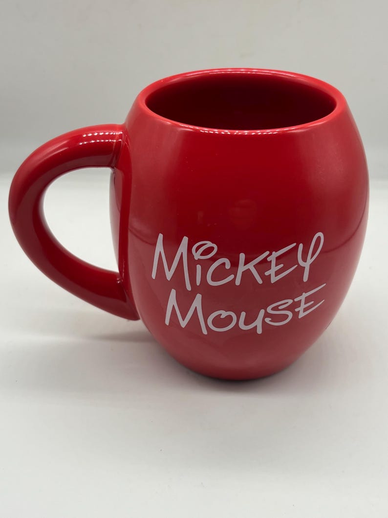 May include: A red ceramic mug with a rounded shape and a large handle. The mug features the words "Mickey Mouse" in white script on the side. The mug is glossy and appears to be new.