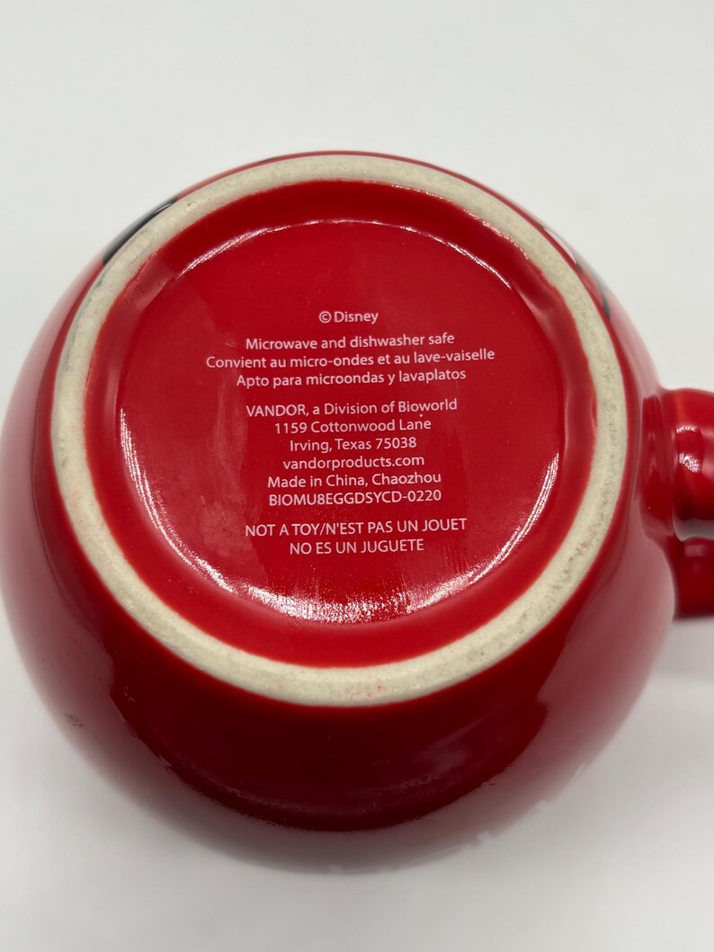 May include: Red ceramic mug with a white rim and handle. The bottom of the mug features text including "Disney", "Microwave and dishwasher safe", and "NOT A TOY/N'EST PAS UN JOUET".