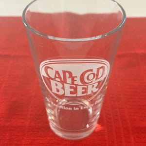 May include: A clear glass beer mug with a white logo that says "Cape Cod Beer" in red lettering. The text below the logo says "A Vacation in Every Pint".