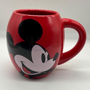 May include: A red ceramic mug featuring a smiling Mickey Mouse design. The mug has a rounded shape with a large handle and a glossy finish. The iconic character's face is prominently displayed on the front.