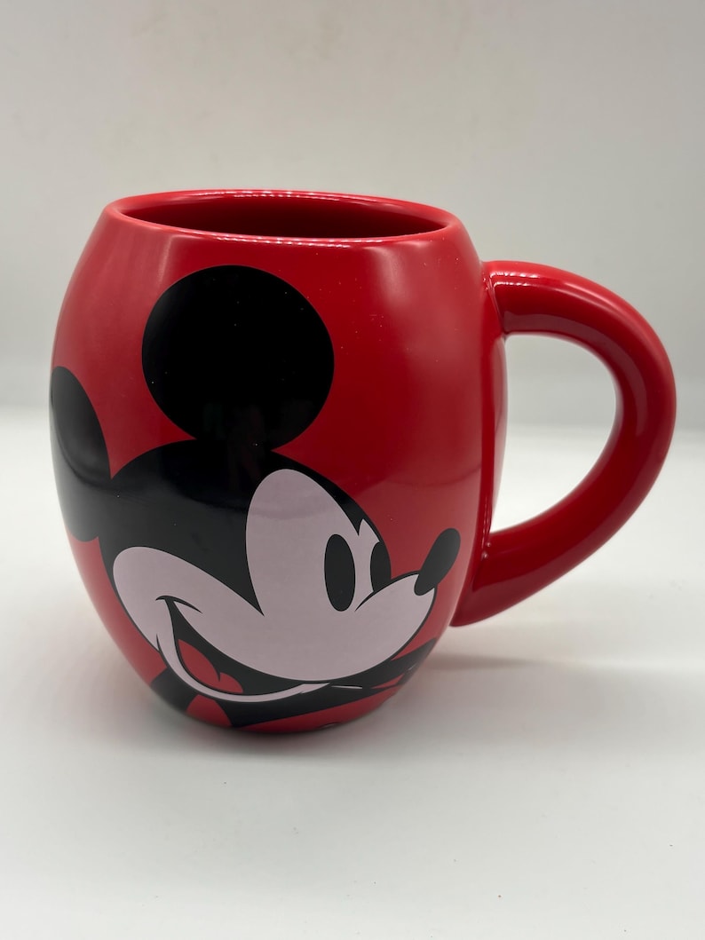 May include: A red ceramic mug featuring a smiling Mickey Mouse design. The mug has a rounded shape with a large handle and a glossy finish. The iconic character's face is prominently displayed on the front.