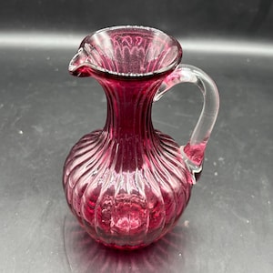 May include: A ruby red glass pitcher with a clear handle. The pitcher has a ribbed design and a spout for pouring. The handle is clear and attached to the side of the pitcher. The pitcher is likely used for serving beverages.