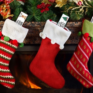 Könnte beinhalten: Drei Weihnachtssocken hängen über einem Kamin. Die Strümpfe sind rot, grün und weiß, mit festlichen Mustern und Bommelakzenten. Jeder Strumpf enthält eine dekorative Karte mit Weihnachtsgrüßen. Der Kamin strahlt ein warmes, einladendes Licht aus.