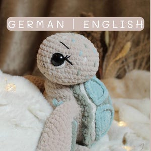 Könnte beinhalten: Gehäkelte Schildkröte aus weichem Beige, Hellblau und Grau. Die Schildkröte hat einen strukturierten Panzer mit blauen Akzenten und ein detailliertes Gesicht mit einem schwarzen Auge. Der Text "GERMAN | ENGLISH" steht oben.