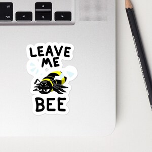 May include: A black and yellow cartoon bee with a grumpy expression. The bee is surrounded by white wings and the text "Leave Me Bee" in black.