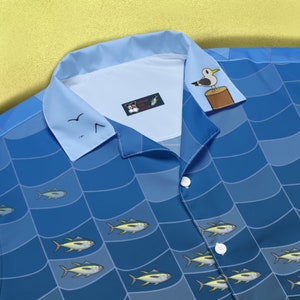 Op de afbeelding: Een blauw-wit gepatroneerd overhemd met knopen, een zeemeeuw-afbeelding en de tekst "Catch the Tuna" op de kraag. Het overhemd heeft verschillende illustraties van tonijn.