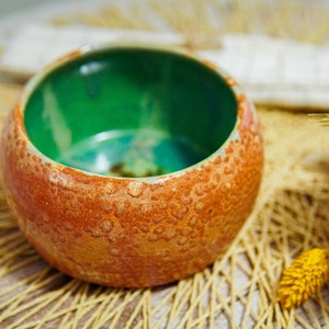 Peut inclure: Un petit bol rond en céramique avec un extérieur orange texturé et un intérieur vert lisse. Le bol est posé sur un tapis tissé.