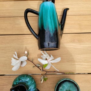 May include: A vintage ceramic tea set with a black and green glaze. The set includes a teapot, creamer and sugar bowl. The teapot has a tall, slender shape with a spout and handle. The creamer and sugar bowl are round with lids. The set is on a wooden surface.