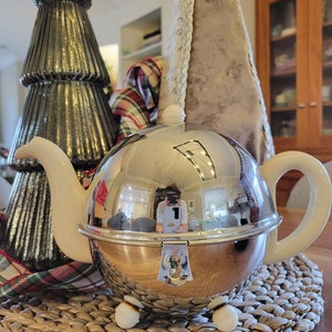 May include: A silver teapot with a white lid and a white handle. The teapot is sitting on a woven placemat. The teapot is reflecting the surrounding environment, including a person holding a phone.