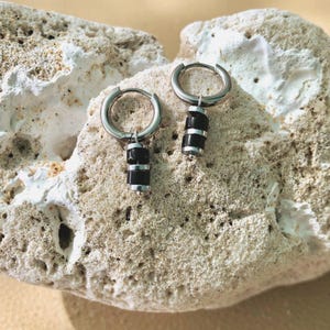 Peut inclure: Une paire de boucles d'oreilles créoles argentées, chacune dotée d'une breloque composée de deux perles cylindriques noires séparées par une fine entretoise en métal argenté. Les boucles présentent une finition métallique polie et un design circulaire.