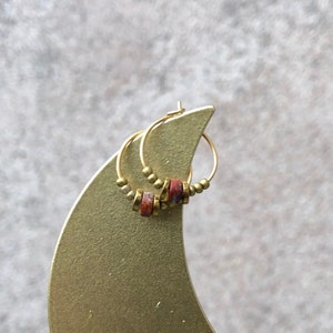 Può includere: Orecchini a cerchio dorati con dettagli in pietra rossa e marrone. Gli orecchini presentano piccole perline dorate e un design circolare. Gli orecchini sono esposti su un supporto color oro su sfondo neutro.
