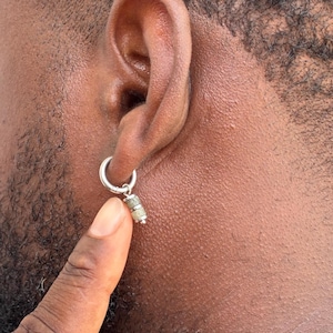 May include: A silver-toned hoop earring featuring a small, cylindrical charm with a textured, mottled grey and beige stone-like inlay. The earring is secured through the earlobe, with the charm hanging vertically from the bottom of the hoop.