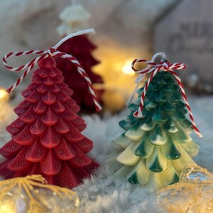 Könnte beinhalten: Nahaufnahme von drei weihnachtsbaumförmigen Kerzen. Die vordere Kerze ist rot mit einer rot-weißen Schnurschleife. Die zweite Kerze ist grün und weiß mit einer Schnurschleife. Die Kerzen stehen auf einer flauschigen weißen Oberfläche.