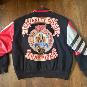 May include: A black varsity jacket featuring red leather sleeves and a large embroidered patch on the back. The patch displays the text Stanley Cup 1997, Detroit Red Wings, and Champions, along with a graphic of the Stanley Cup trophy.