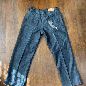 May include: Dark wash denim jeans with a straight leg cut. The back right pocket features a white embroidered wing design. The jeans have a classic five-pocket design and a brown leather patch on the waistband.