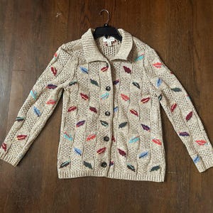 May include: Beige knit cardigan with a collared neckline and button-down front. The sweater features a pattern of colorful leaf-shaped designs in red, blue, green, and purple. The long-sleeved cardigan is hanging on a black hanger.