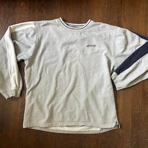 May include: A light gray long-sleeve sweatshirt featuring a crew neckline with navy and white striped trim. The garment displays the text CHAPS embroidered in small letters on the chest, with a white stripe on the right sleeve and a navy stripe on the left.
