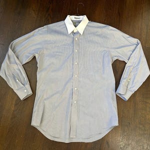 May include: A long-sleeve button-down shirt in a light blue color featuring a solid white pointed collar. The shirt includes a single chest pocket on the left side, a front button placket, and buttoned cuffs on each sleeve, presented on a hanger.