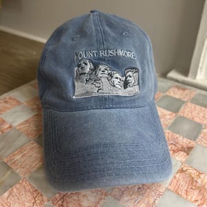 May include: A blue baseball cap with the embroidered text "MOUNT RUSHMORE" above an image of the Mount Rushmore presidents. The cap is made of a soft fabric and has a curved brim.