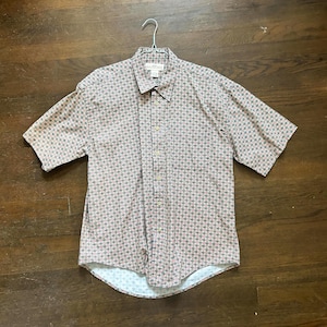 May include: Short-sleeved button-down shirt with a repeating pattern of small red and green shapes on a light brown background. The shirt is hanging on a metal hanger against a dark wood surface.
