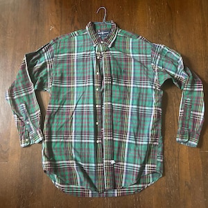 May include: Green, red, and white plaid button-down shirt. The long-sleeved shirt is hanging on a hanger. The shirt has a button-down collar and a small logo on the chest. The plaid pattern is a classic design.