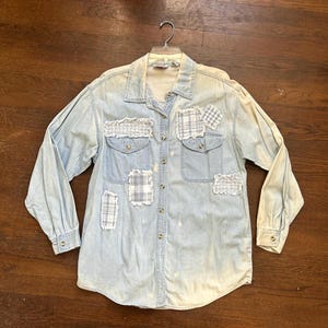 May include: Light blue denim shirt with long sleeves and a collared neckline. The shirt features decorative patches of plaid fabric and fringed detailing on the pockets. The shirt is button-down and has two chest pockets.
