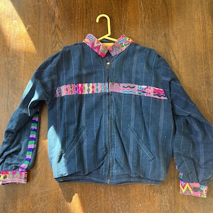 May include: A dark blue zip-up jacket with colorful embroidered details. The jacket features a striped pattern, a vibrant embroidered band across the chest, and cuffs with matching embroidery. The jacket is hanging on a yellow hanger.