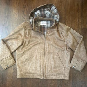 May include: A tan zip-up jacket with a hood, featuring a brown, white, and tan plaid lining. The jacket has long sleeves with buttoned cuffs, two front pockets, and a label inside the collar that reads "Made in USA" with additional text.