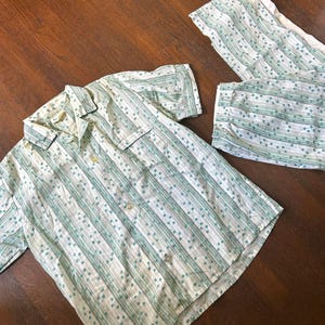 May include: A two-piece pajama set featuring a short-sleeved button-down shirt and matching pants. The set is white with vertical stripes and small teal dots. The shirt has a collar and two chest pockets.