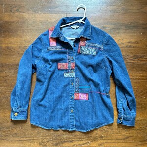 May include: A blue denim button-down shirt with long sleeves. The shirt features colorful patchwork designs in shades of pink, red, and blue. The shirt is hanging on a white hanger.