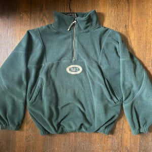 May include: A dark forest green fleece pullover featuring a quarter-zip neckline with a silver-toned zipper. The garment includes a central oval patch embroidered with the text PAR 71, long sleeves with elastic cuffs, and a drawstring hem.