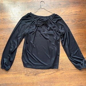 May include: Black long-sleeve blouse with a round neckline and a tie detail at the front. The blouse has a silky texture and is hanging on a metal hanger. The sleeves are long and end in a gathered cuff.