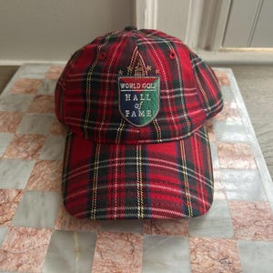 May include: A red, green, and black plaid baseball cap with the "World Golf Hall of Fame" emblem. The cap has a curved brim and a structured crown. The emblem is a patch with the text "World Golf Hall of Fame".