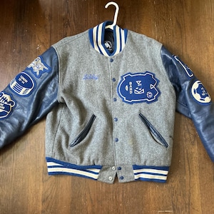 May include: A grey wool varsity jacket with blue leather sleeves, featuring blue and white striped ribbed cuffs, collar, and hem. The jacket has blue patches on the sleeves and chest, including text reading "BAND", "TENOR 12-14", and "DRUM CAPTAIN".