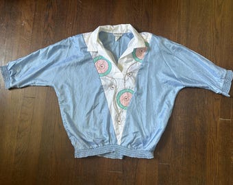 May include: A light blue blouse with a white V-shaped front panel featuring pink and teal circular embroidery. The garment has a white collar, three-quarter length sleeves with gathered cuffs, and a gathered elasticized waistband at the bottom.