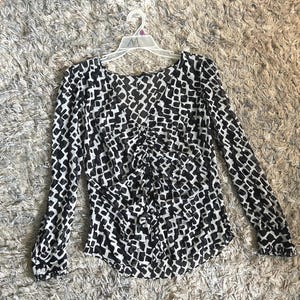 May include: Black and white patterned long-sleeve blouse. The blouse features a ruched front detail and is hanging on a white hanger. The pattern consists of small, irregular black shapes on a white background.
