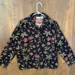May include: A black long-sleeve button-up jacket featuring a woven floral pattern in shades of red, pink, and green. The garment has a standard collar, a straight hem, and is displayed on a white plastic hanger against a wooden background.