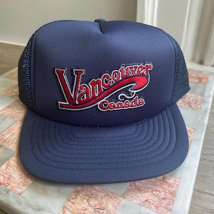 May include: A navy blue trucker hat featuring a mesh back and a solid front panel. The front displays an embroidered patch with the text Vancouver in red and white script, with the word Canada in smaller red and white lettering underneath.
