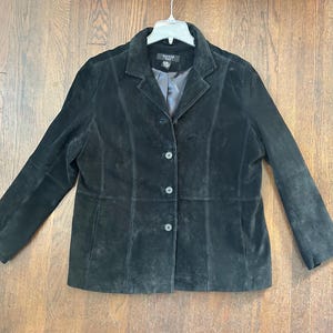 May include: A black suede blazer featuring a notched lapel, long sleeves, and a front button closure with three visible buttons. The jacket includes vertical seam detailing along the front panels and a dark interior lining visible at the collar.