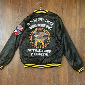 May include: A black satin bomber jacket with yellow and black striped ribbed trim. The back features embroidered text reading 217TH MILITARY POLICE ALABAMA NATIONAL GUARD, PRATTVILLE ALABAMA PHILIPPINES '86, and a central United States Army eagle emblem.