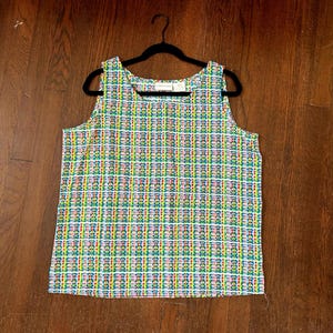 May include: A sleeveless top with a colorful plaid pattern in shades of green, yellow, red, and white. The top is hanging on a black hanger against a dark wood background. The fabric has a textured appearance.