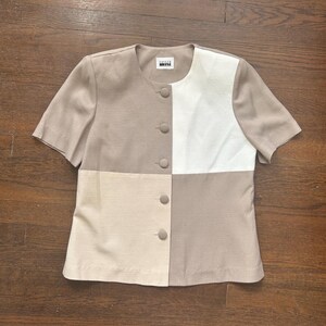 May include: A short-sleeved, button-down blouse in a color-block design. The top features a beige and white color scheme, with a square of white fabric on the right side. The blouse has a round neckline and a small label at the top.