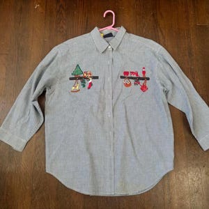 May include: A long-sleeved button-down shirt with a blue and white checkered pattern. The shirt features embroidered Christmas-themed designs, including a Christmas tree, stockings, and ornaments. The shirt is hanging on a pink hanger.