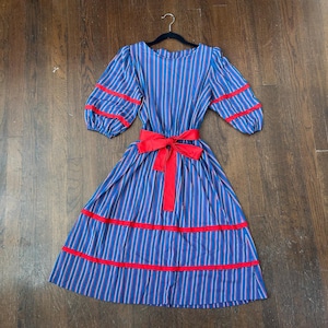 May include: Blue and red striped dress with a red bow and trim. The dress has short, puffy sleeves and is hanging on a black hanger. The dress is made of a lightweight fabric and has a vintage style.