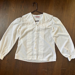 May include: A white long-sleeved button-down blouse featuring a large sailor-style collar with white lace trim, vertical pintuck pleats along the front placket, and gathered cuffs at the wrists. The shirt includes small white buttons down the center.