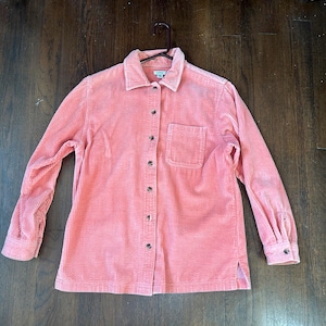 May include: A coral-colored corduroy shirt with long sleeves, a button-down front, and a single chest pocket. The shirt is hanging on a black hanger against a dark wood background. The shirt has a classic collar and buttoned cuffs.