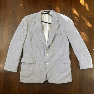 May include: A seersucker blazer with a blue and white striped pattern. The jacket has a notched lapel, two front pockets, and white buttons. It is hanging on a black hanger against a dark wood background.