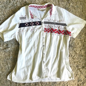 May include: White short-sleeved button-down shirt with decorative embroidered patterns in black, red, and navy blue. The collar and upper chest feature a plaid design. The shirt has a textured fabric and a relaxed fit.
